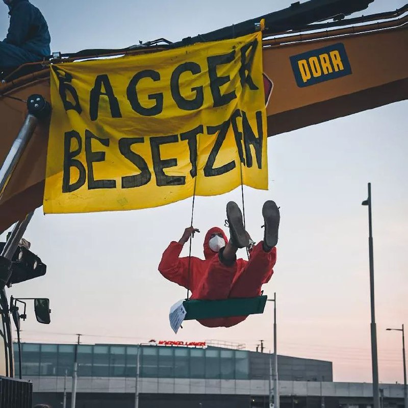 Aktivist schaukelt auf besetztem Bagger, von dem ein Banner h&auml;ngt